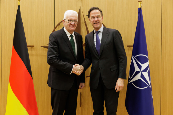 Ministerpräsident Winfried Kretschmann (links) und NATO-Generalsekretär Mark Rutte (rechts) stehen zwischen Fahnen und geben sich die Hand.