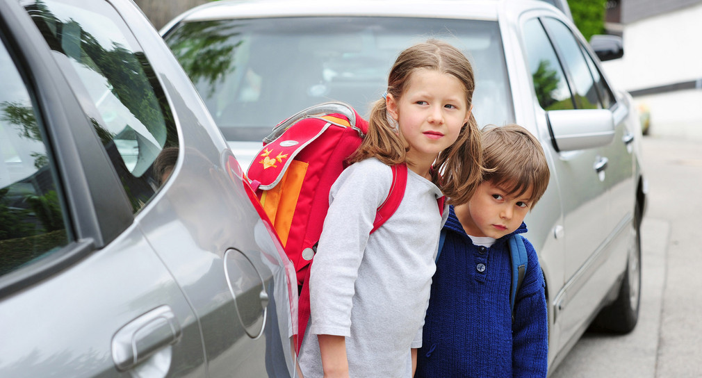 Zwei Kinder mit Schultaschen zwischen geparkten Autos am Straßenrand. Quelle: Fotolia