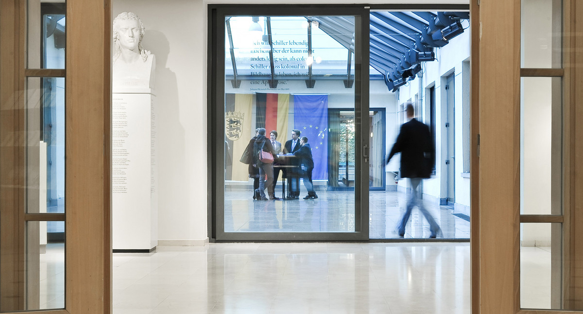 Eingangssituation mit Blick in den glasüberdachten Innenhof mit Gruppe von Menschen vor Fahnen.