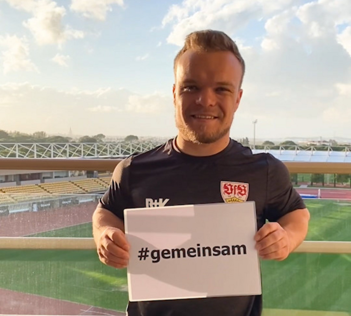 Der Sportler Niko Kappel hält ein weißes Blatt mit der Aufschrift #gemeinsam in die Kamera. Er steht im VFB Stadion bei blauem Himmel. 