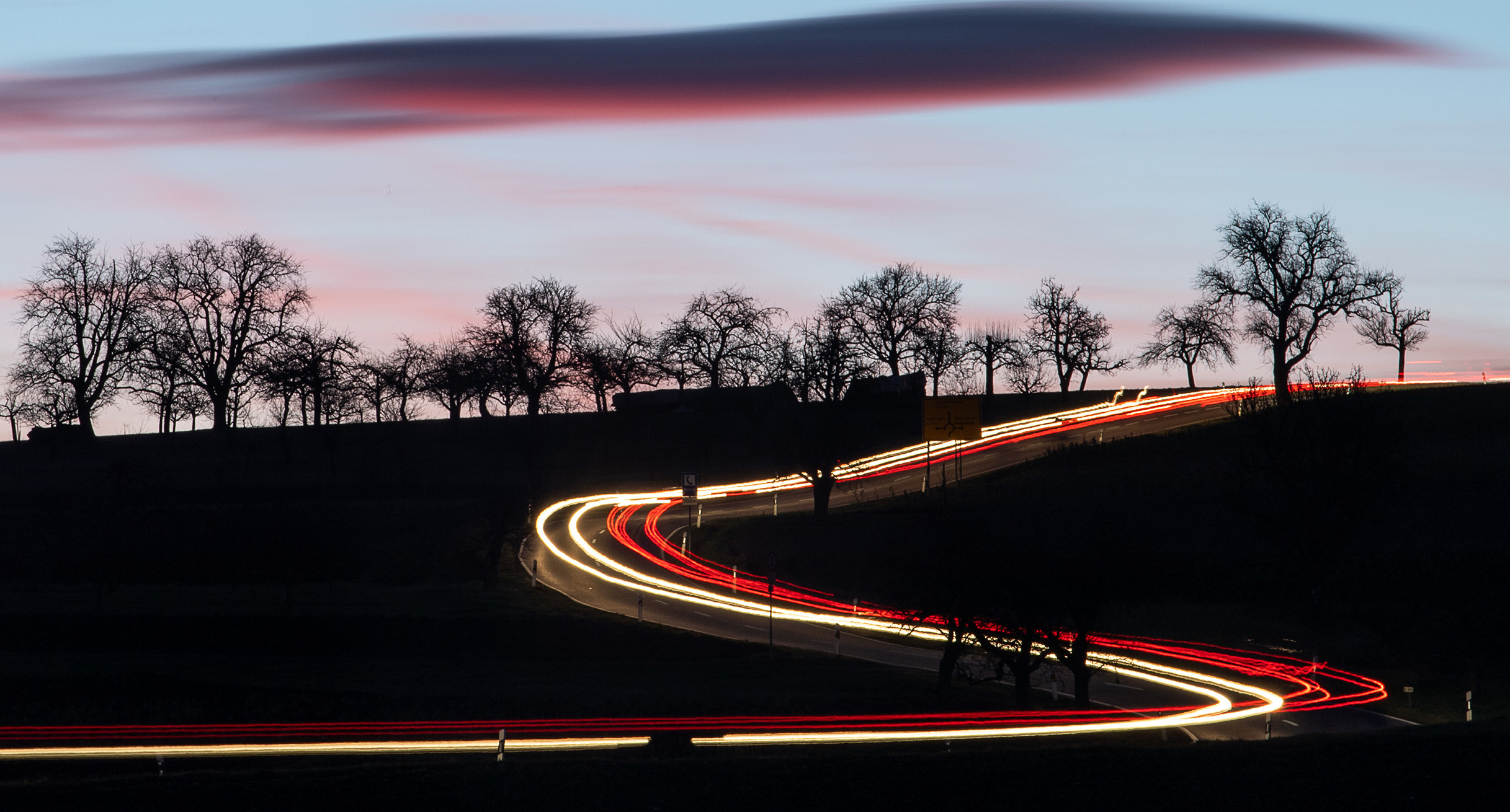 Autos fahren am frühen Morgen auf einer Landstraße bei Vaihingen an der Enz und hinterlassen eine Leuchtspur auf dem Foto.']
