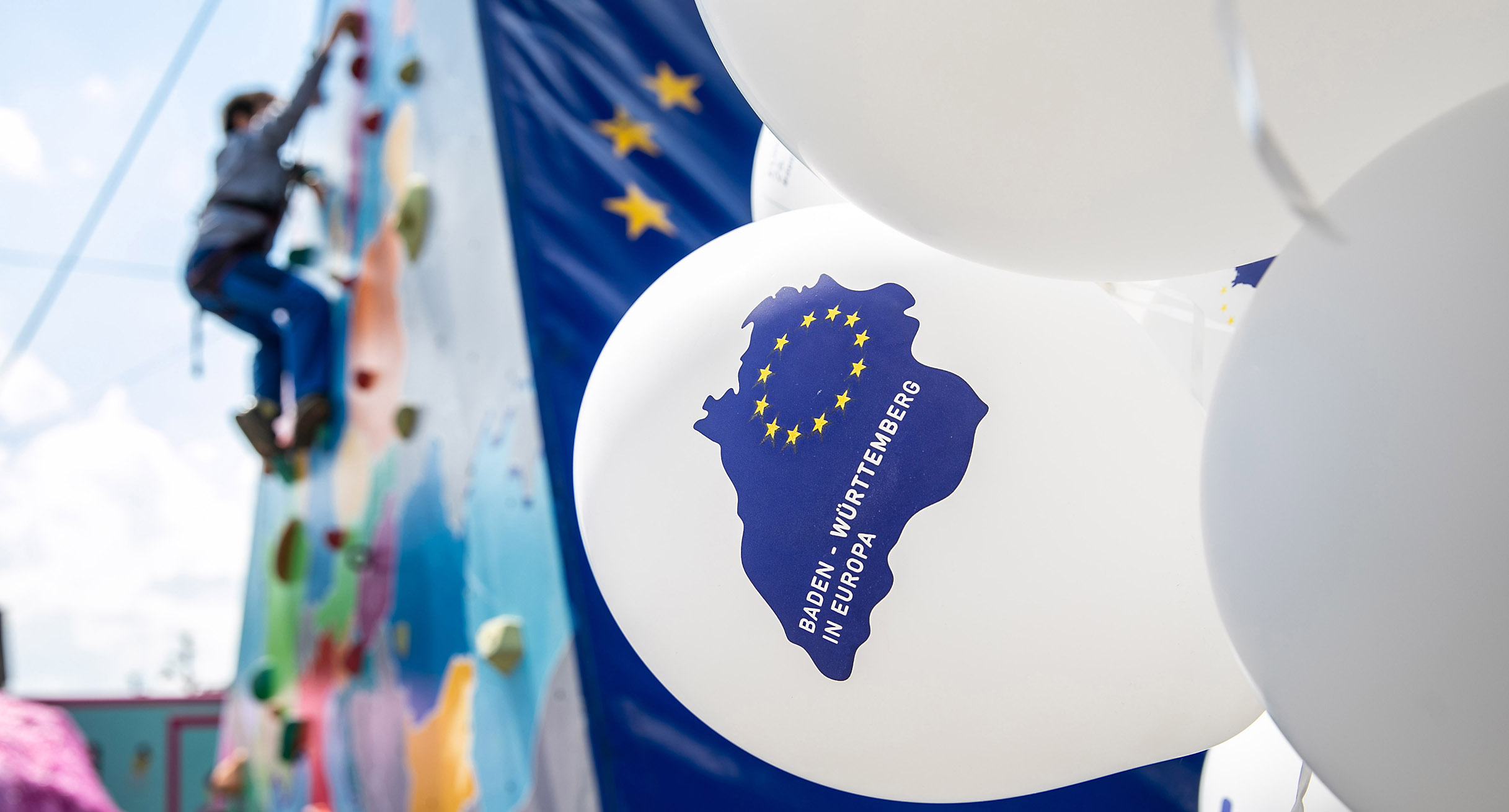 Einige wei&szlig;e Luftballons mit der Aufschrift &bdquo;Baden-W&uuml;rttemberg in Europa&ldquo; sind im Vordergrund zu sehen, im Hintergrund sind die EU-Flagge und eine Person an einer Kletterwand zu erkennen.